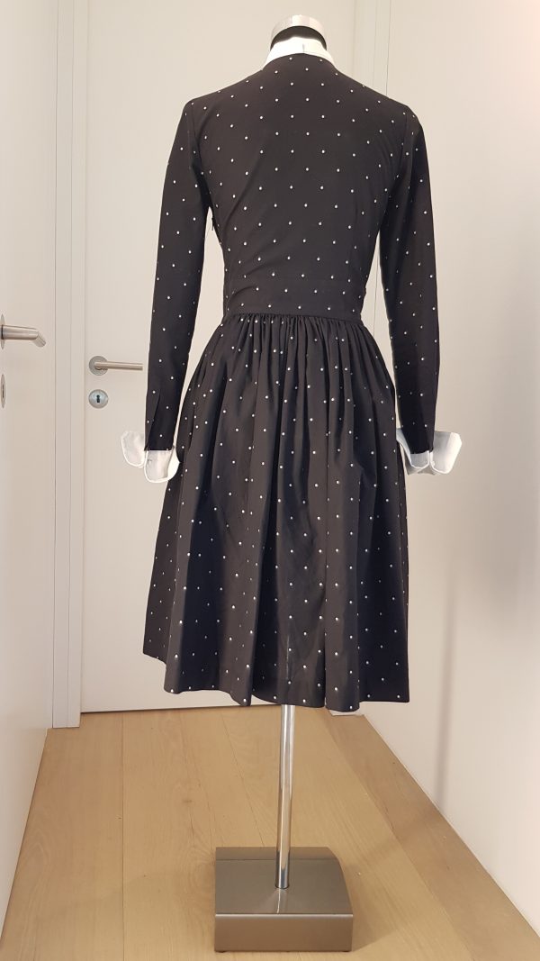 Raffinierts Smokingkleid mit leicht ausgestelltem Rock, mit versteifter Smokingbrust in Piqué, weißen Knöpfen, weißem Mascherl in Piqué, weiße Manschetten mit Manschettenknöpfen, schwarzer Baumwollstoff mit kleinen weißen Tupfen, seitlicher Zippverschluss, breiter Bund